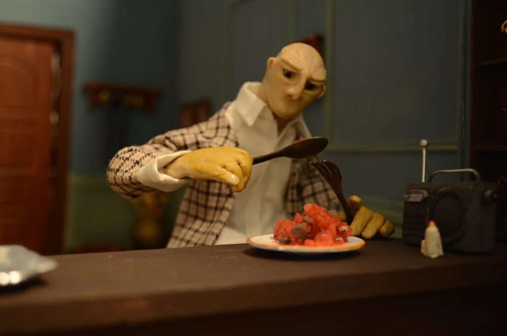 Stop Motion Figur eines Mannes sitzt am Küchentresen und isst von seinem Teller - im Hintergrund sehen wir die Haustür und das Wohnzimmer